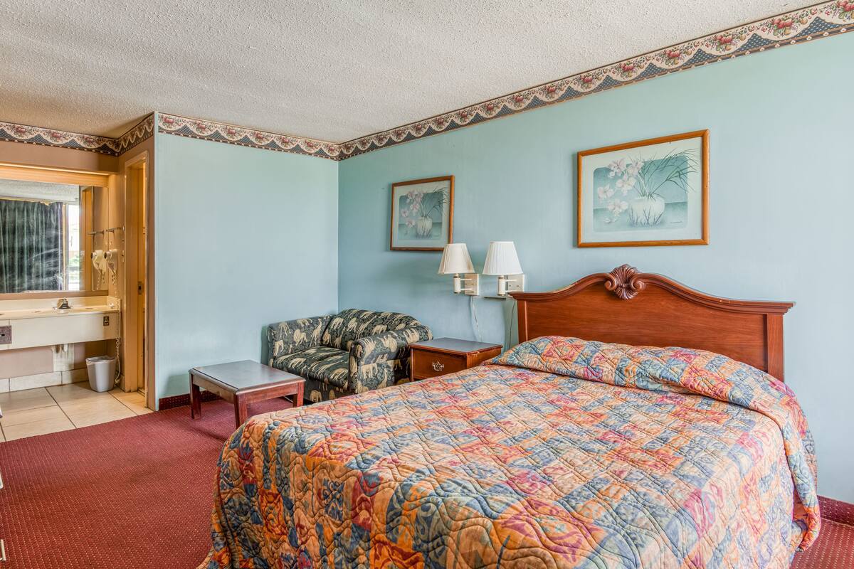 A hotel room with a bed, a chair, a desk, and two wall-mounted lamps.