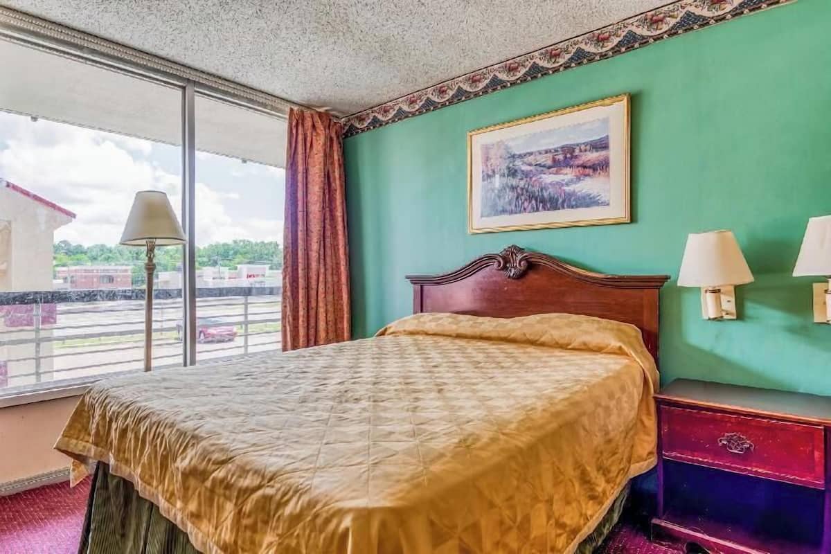 A hotel room with a large bed, a painting on the wall, and a view of the outside.
