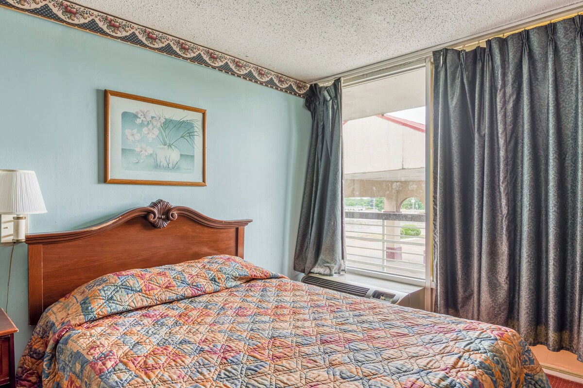 Una habitación de hotel con una cama, un cuadro en la pared y una ventana con cortinas.