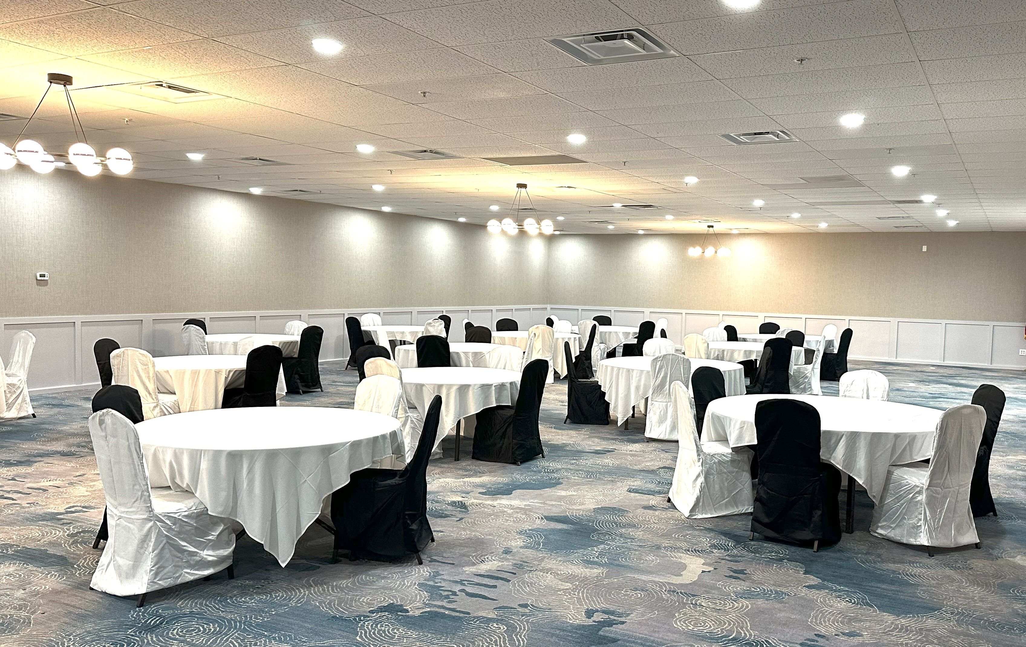 A large conference room with multiple round tables, each with a white tablecloth and black chairs. The room has a high ceiling with recessed lighting and a patterned carpet.
