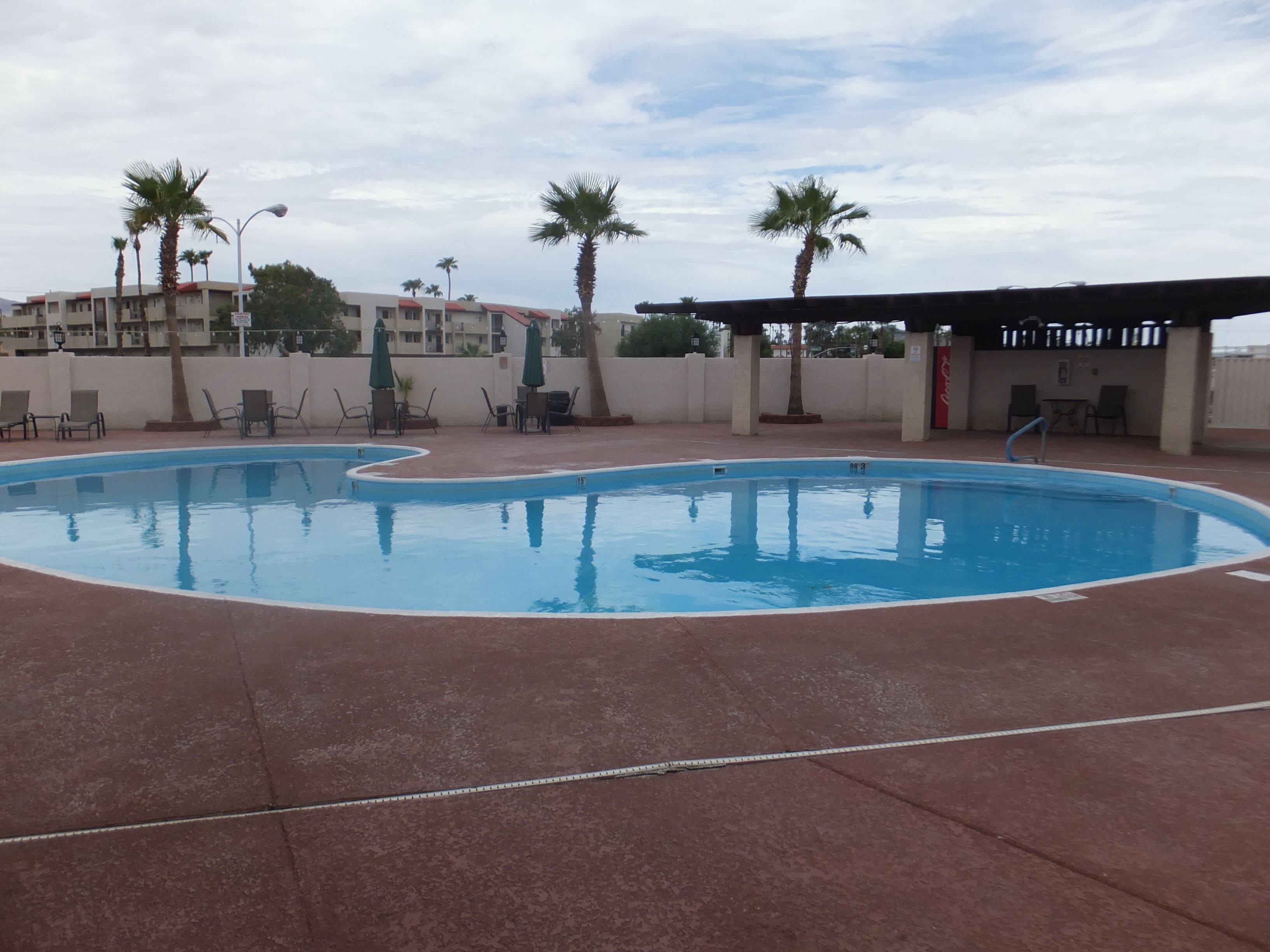 Piscina con sillas de descanso y sombrillas, rodeada de palmeras y una zona cubierta con asientos.