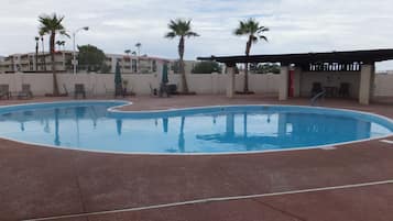 Piscina con sillas de descanso y sombrillas, rodeada de palmeras y una zona cubierta con asientos.