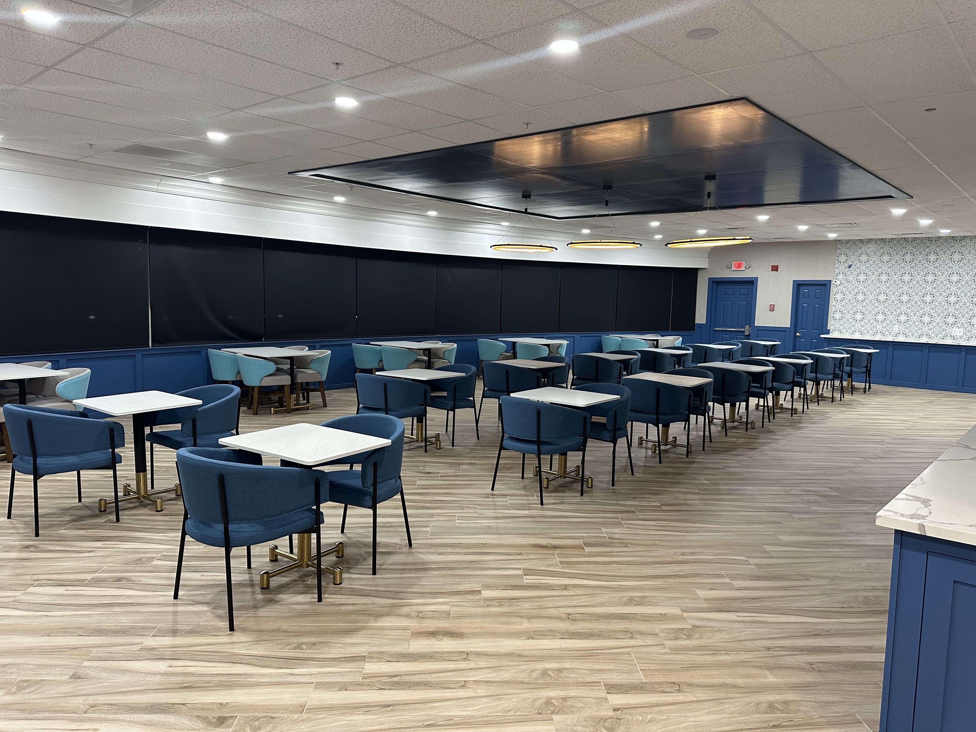 Un vaste espace intérieur aménagé en salle à manger, avec des rangées de tables et de chaises, une scène et un mur décoré de motifs.