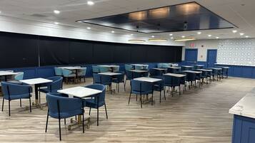 Un vaste espace intérieur aménagé en salle à manger, avec des rangées de tables et de chaises, une scène et un mur décoré de motifs.