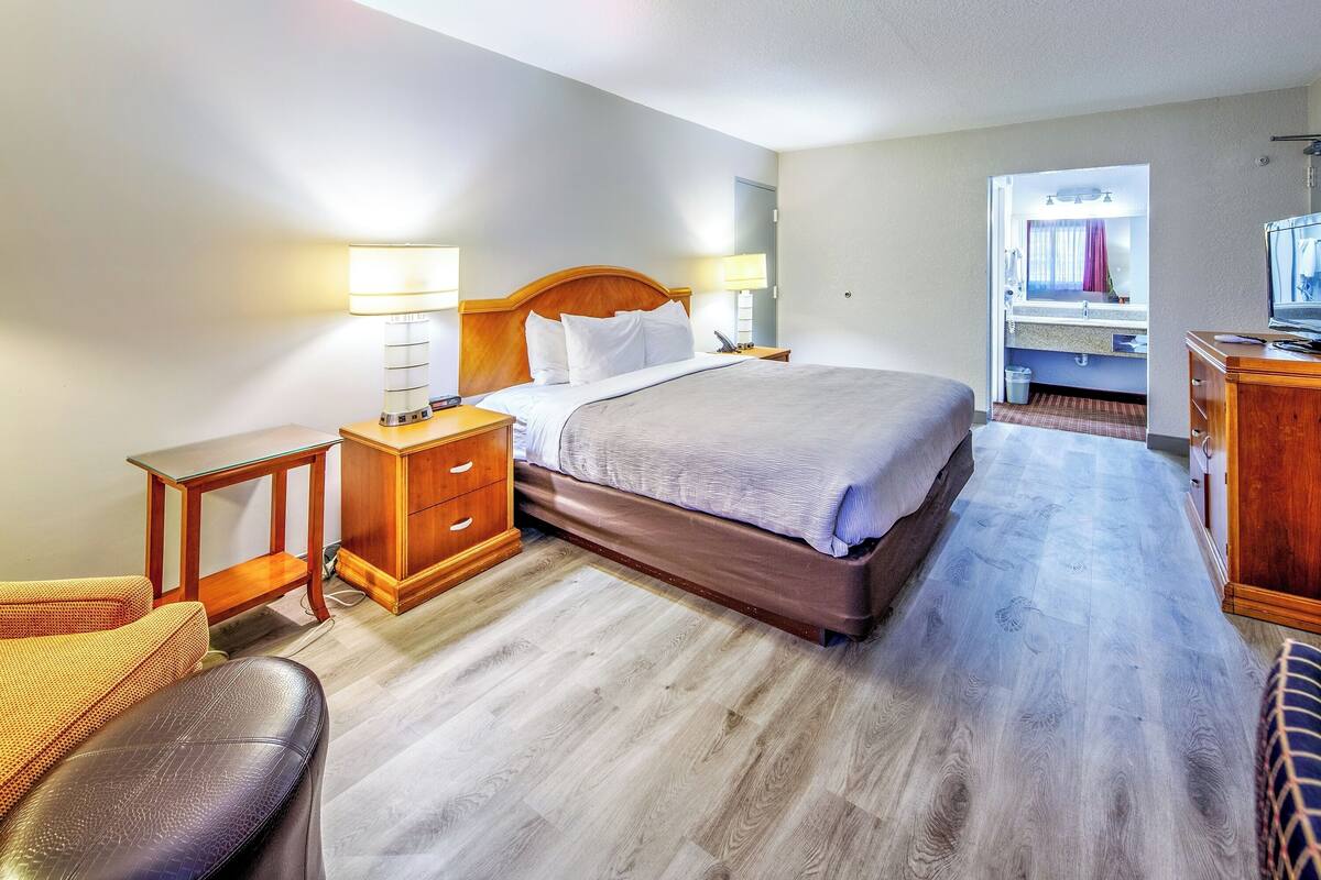 A hotel room with a bed, bedside tables, a wooden dresser, a television, and a bathroom visible through an open door.