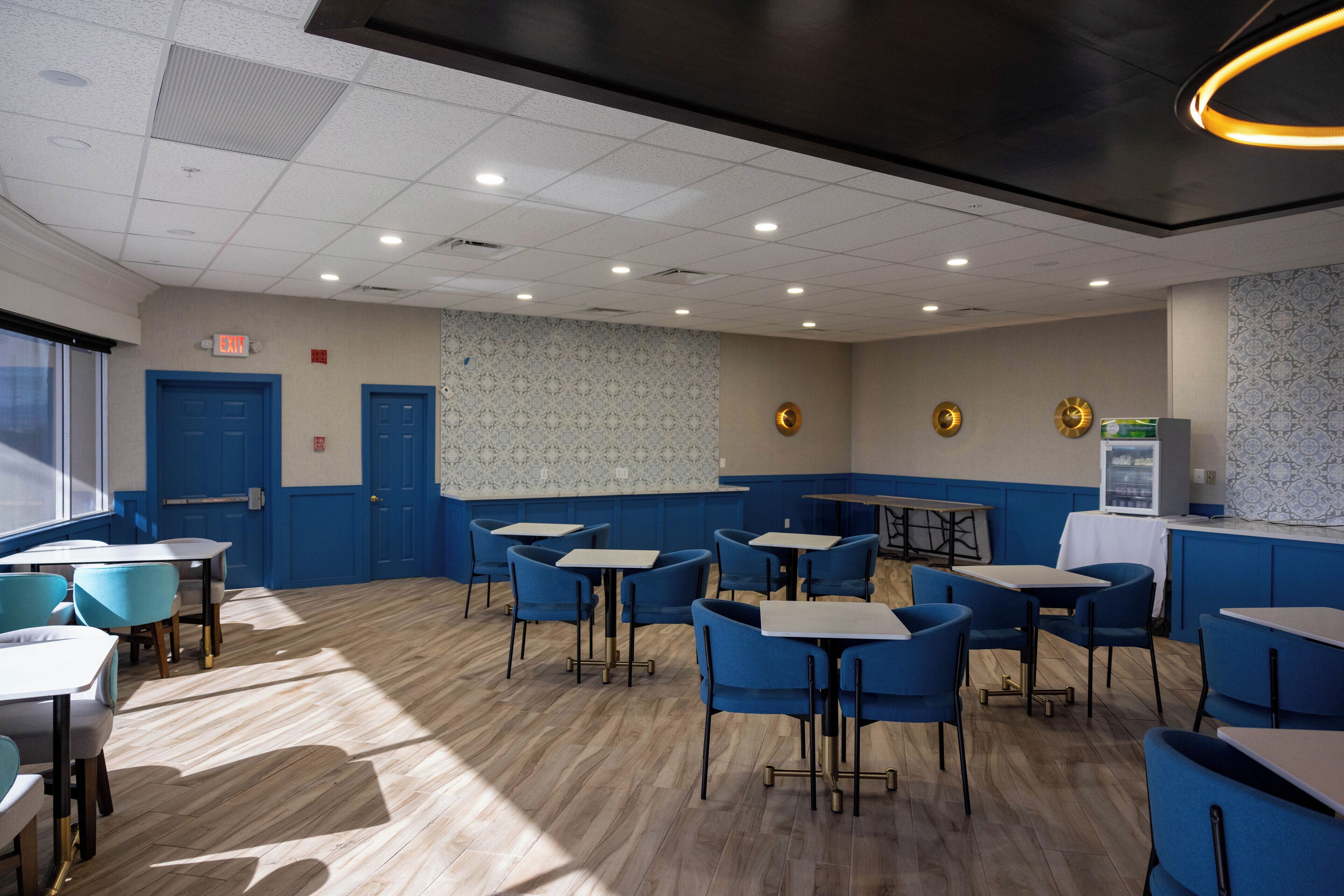 L’intérieur d’un restaurant, avec des chaises et des tables bleues, un papier peint à motifs et une horloge au mur.
