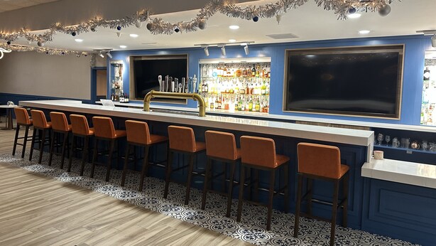 A bar area with a long counter, multiple stools, and a variety of bottles and glasses on shelves.