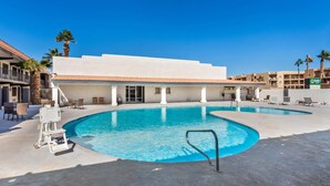 A swimming pool with lounge chairs and a lifeguard chair, surrounded by buildings with a tiled roof and palm trees.