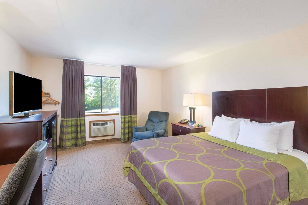 Ein Hotelzimmer mit einem Bett, einem Schreibtisch mit Fernseher, einem Sessel, einer Lampe und einem Fenster mit Vorhängen.