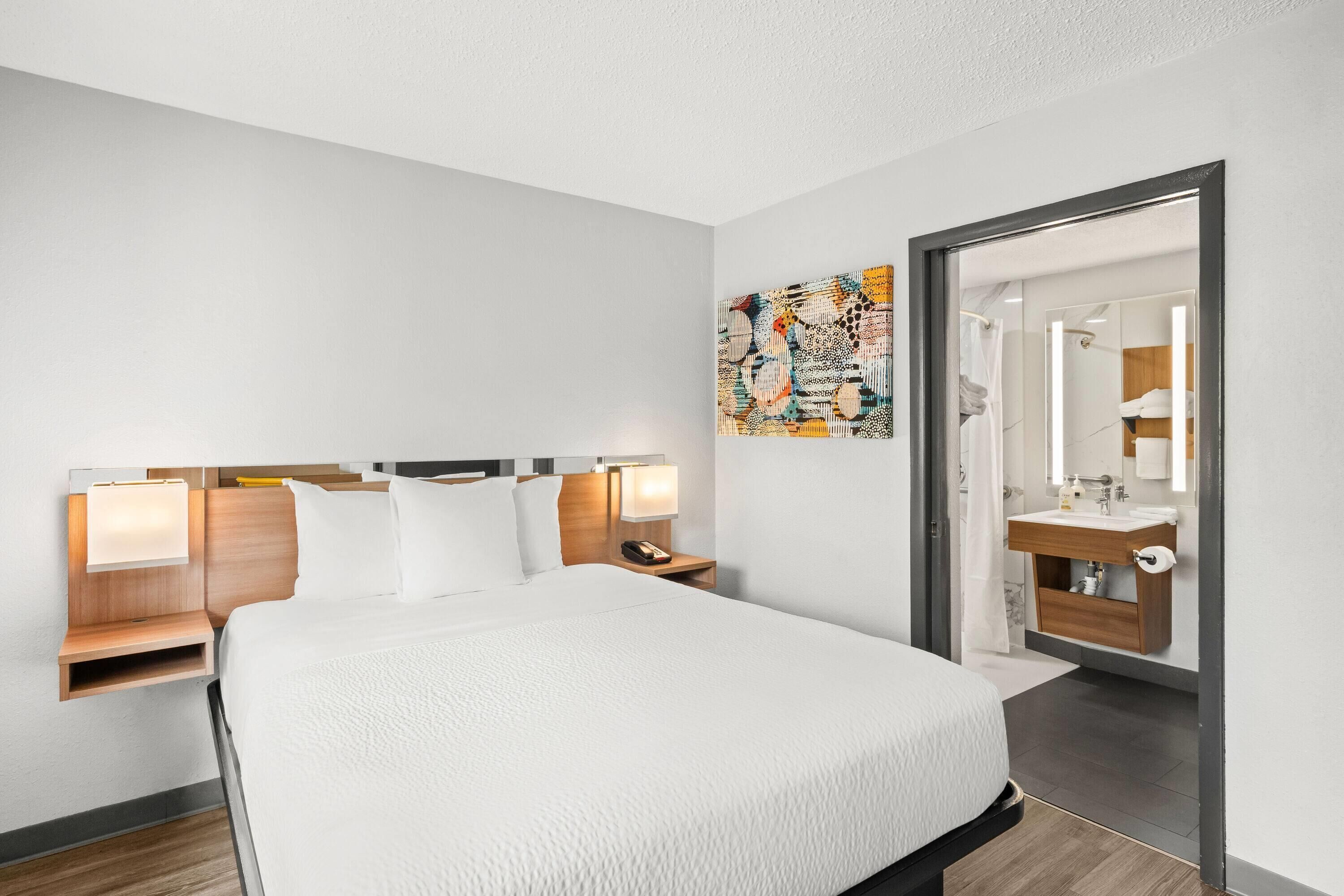 A modern hotel room with a large bed, bedside tables with lamps, a bathroom visible through an open door, and a colorful abstract painting on the wall.