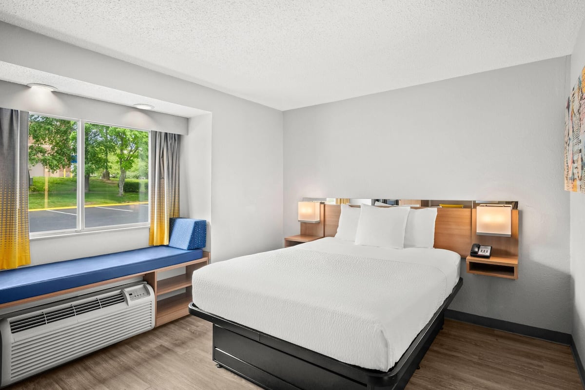 A hotel room with a large bed, a bench by the window, a bedside table with a lamp, and a wall-mounted phone.