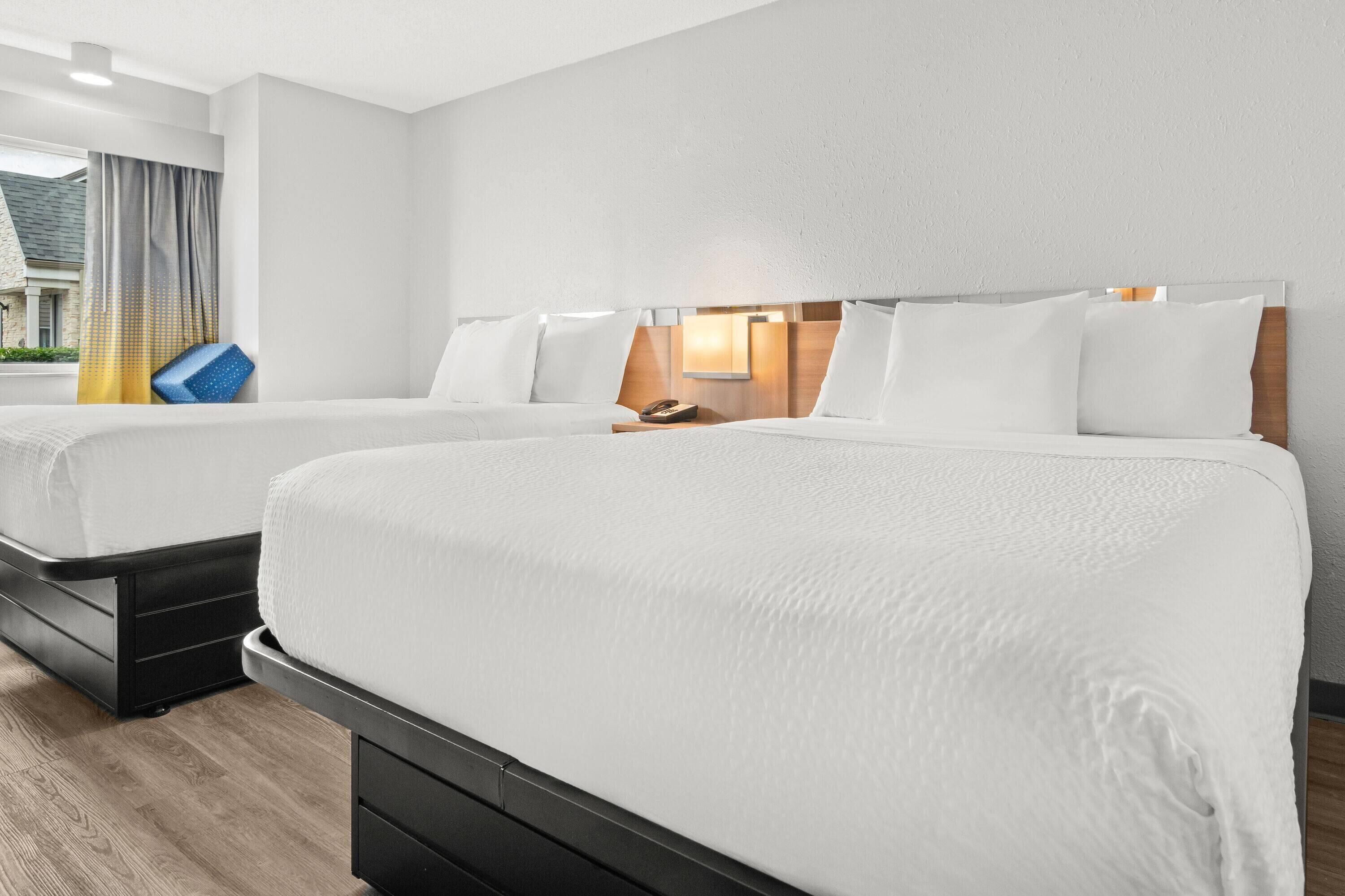 Two beds with white linens, a wooden headboard, a nightstand, and a window with curtains.