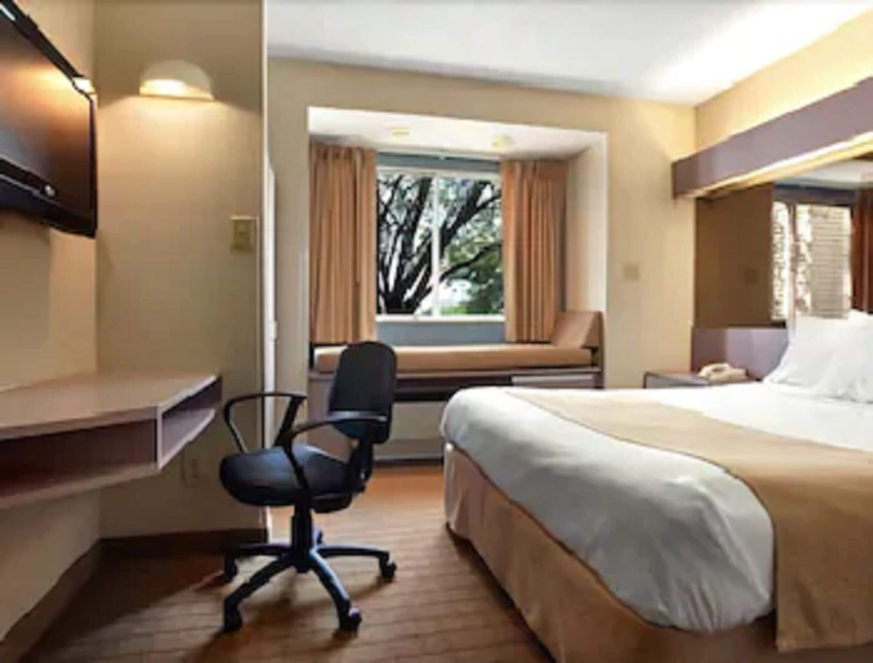 A hotel room with a bed, a desk, a chair, a window with curtains, and a telephone.