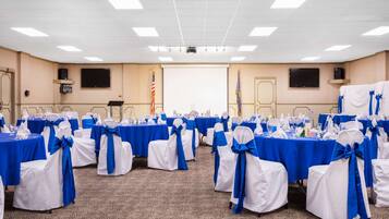Une salle de conférence avec des rangées de tables et de chaises, chacune ornée d’un ruban bleu, et un écran de projection à l’avant.