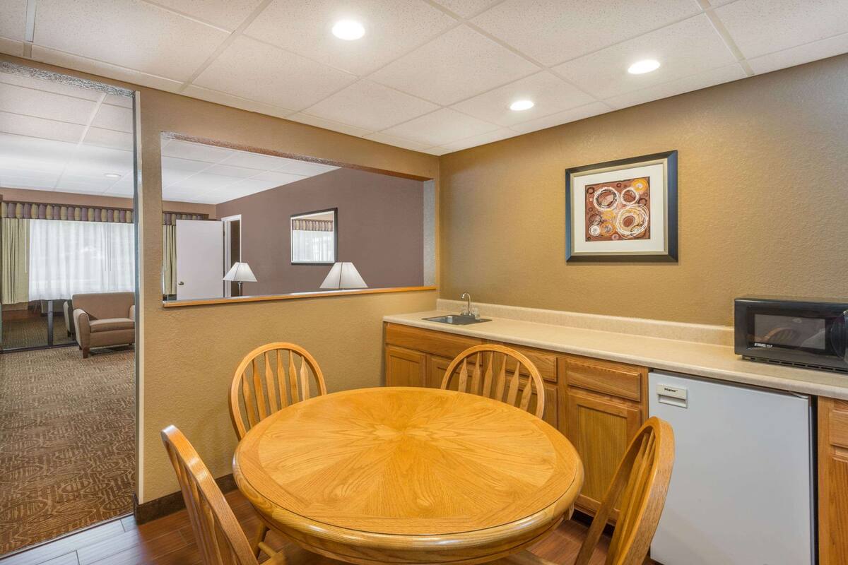 A compact hotel room with a wooden dining table, four chairs, a small kitchenette, a microwave, and a framed artwork on the wall.