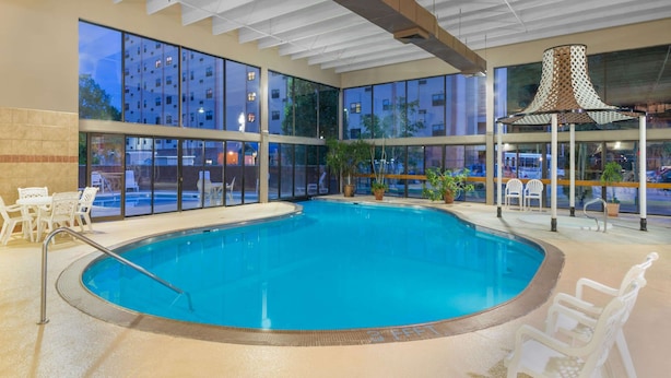 An indoor swimming pool with clear glass walls, white lounge chairs, and potted plants. - Ramada Hotel & Conference Center by Wyndham Paintsville (Paintsville)