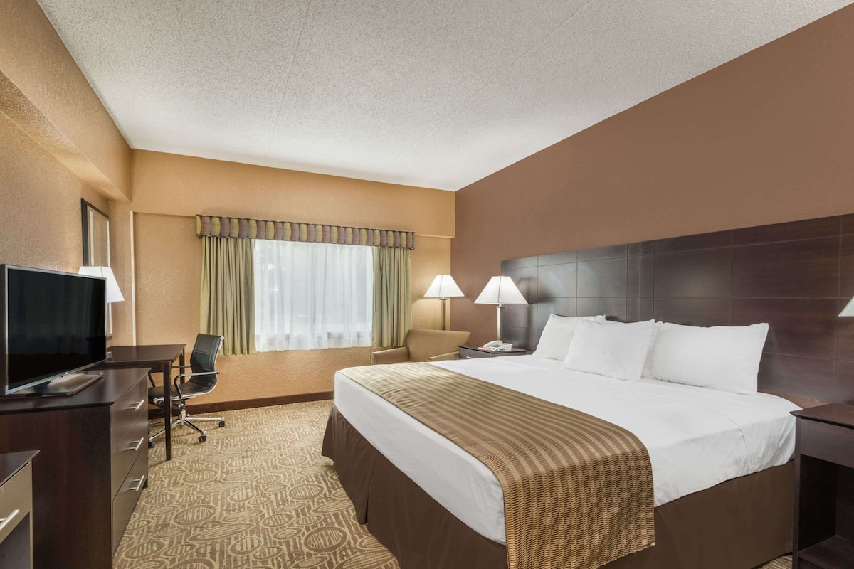A hotel room with a large bed, a desk with a television, two bedside lamps, a chair, and a window with curtains.