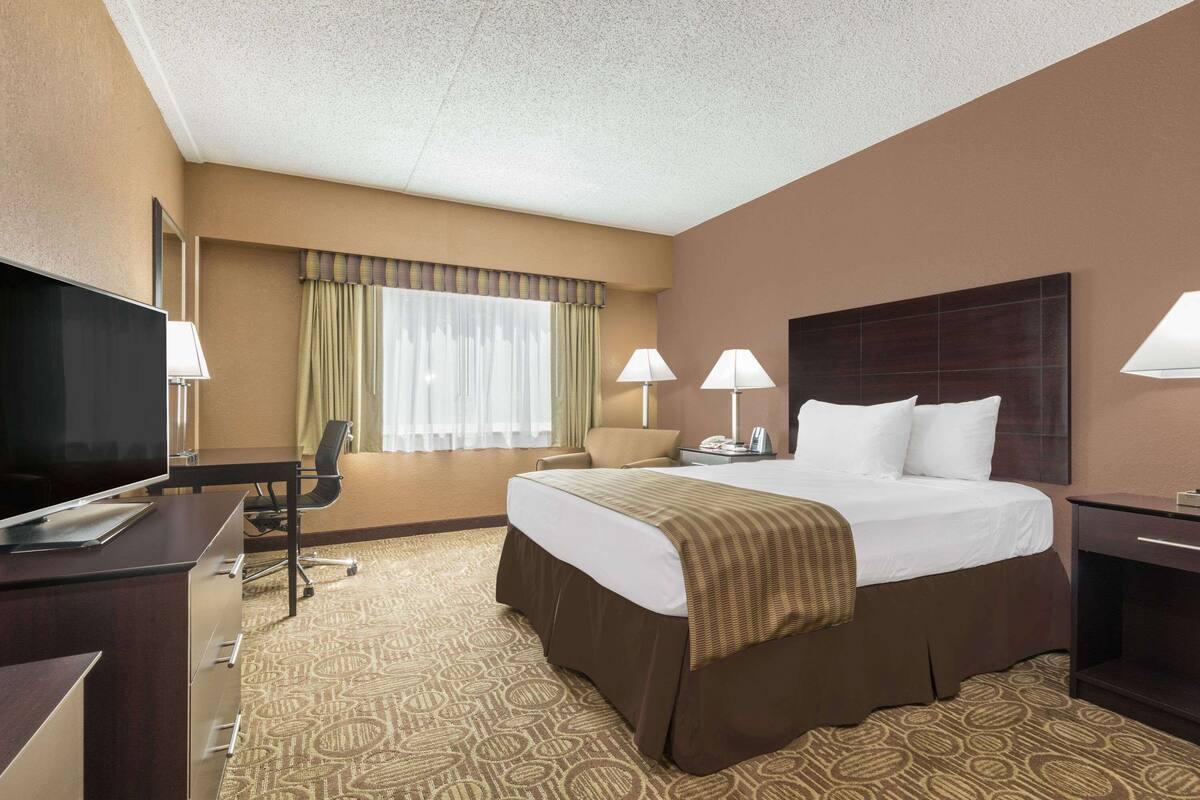 A hotel room with a large bed, a desk with a TV, two lamps, and a window with curtains.