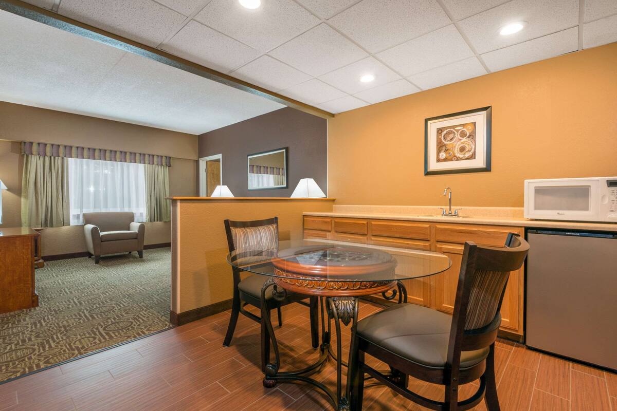 A hotel room with a dining area featuring a glass-top table, two chairs, a sofa, a kitchen area with a microwave, and a refrigerator.