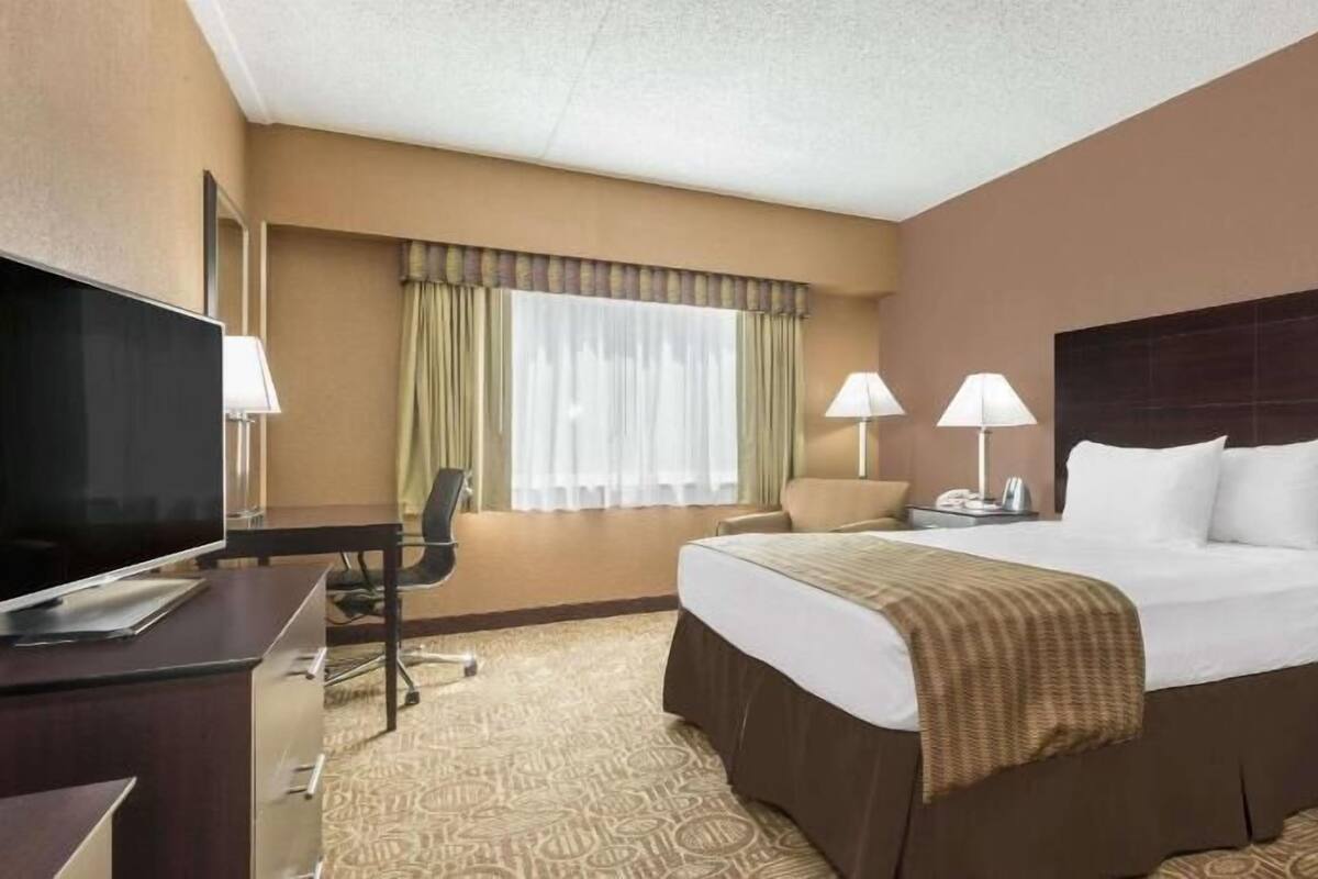 A hotel room with a bed, desk, chair, TV, lamps, and a window with curtains.