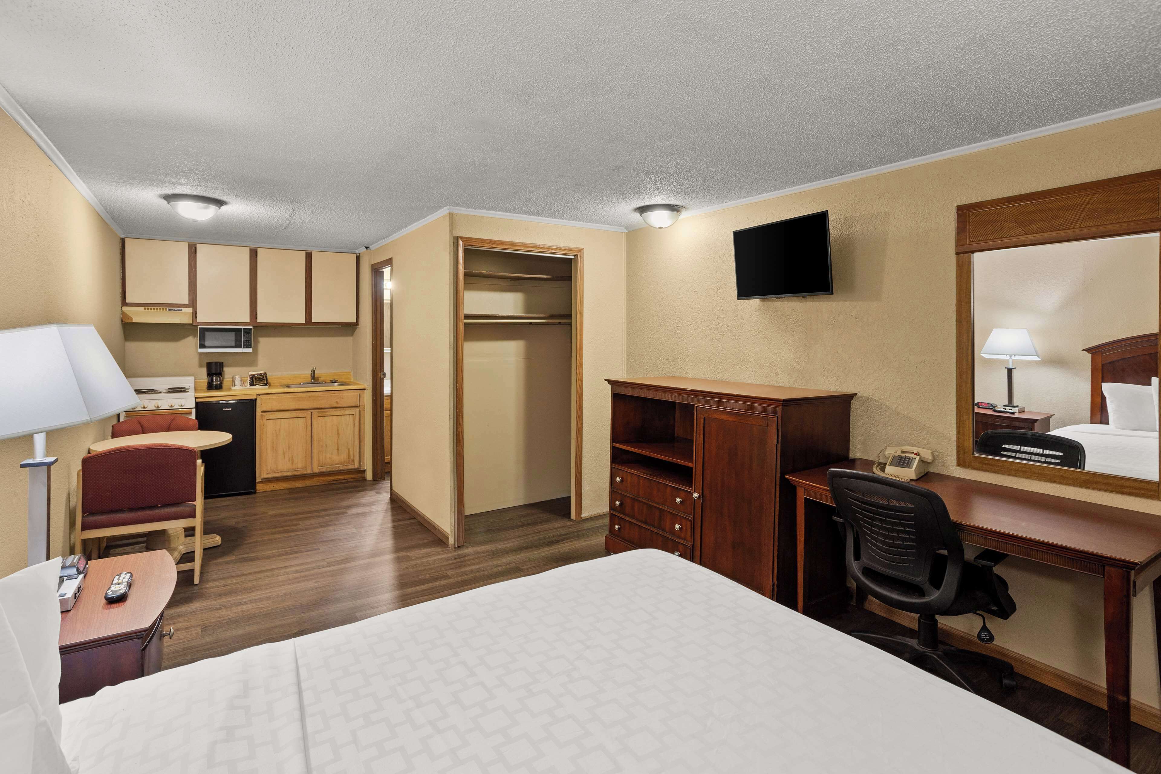 Quarto de hotel com cama, secretária, cadeira, kitchenette e televisão presa na parede.
