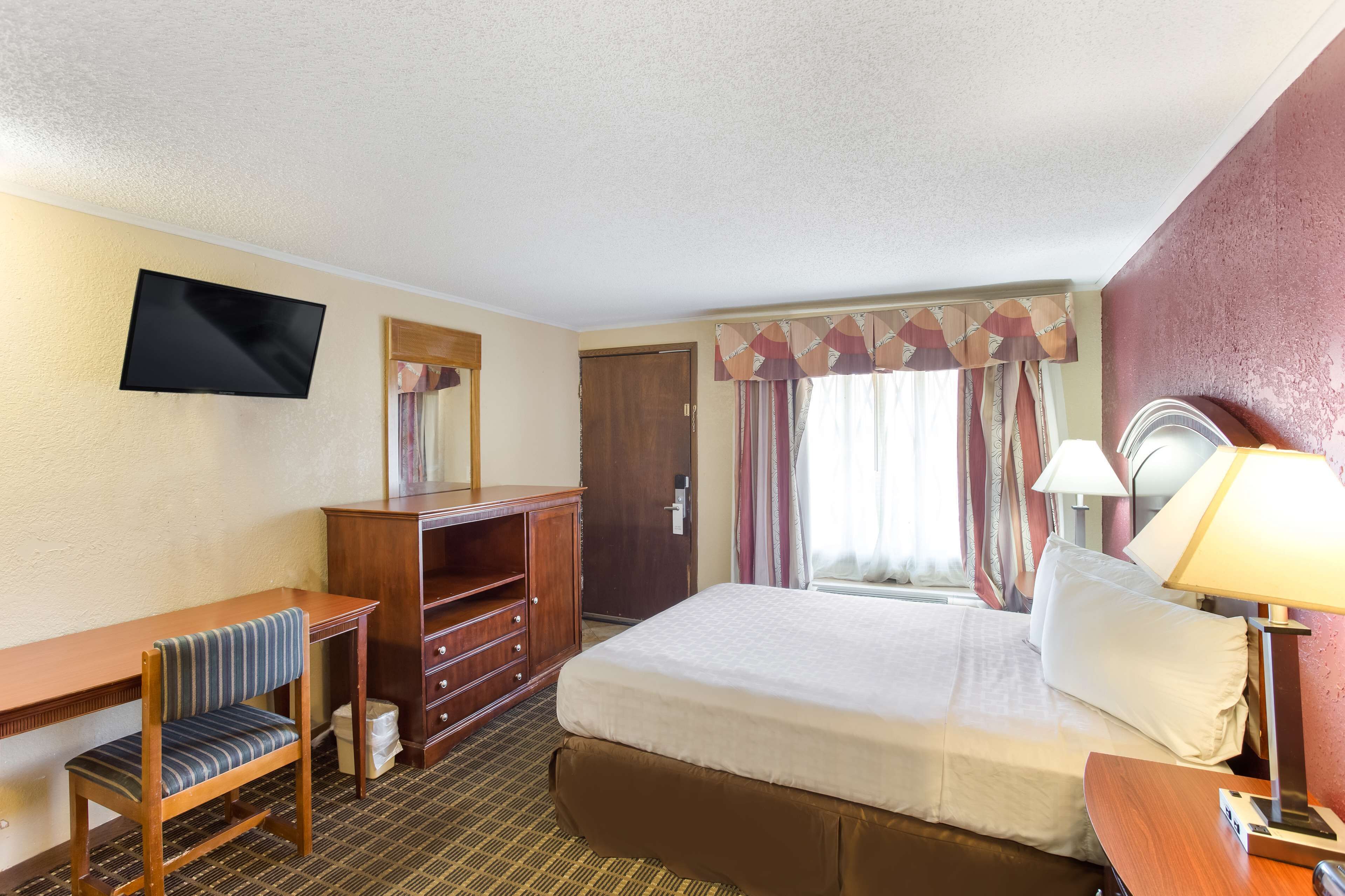 Habitación de hotel con cama, escritorio, silla, televisor, armario y ventana con cortinas.