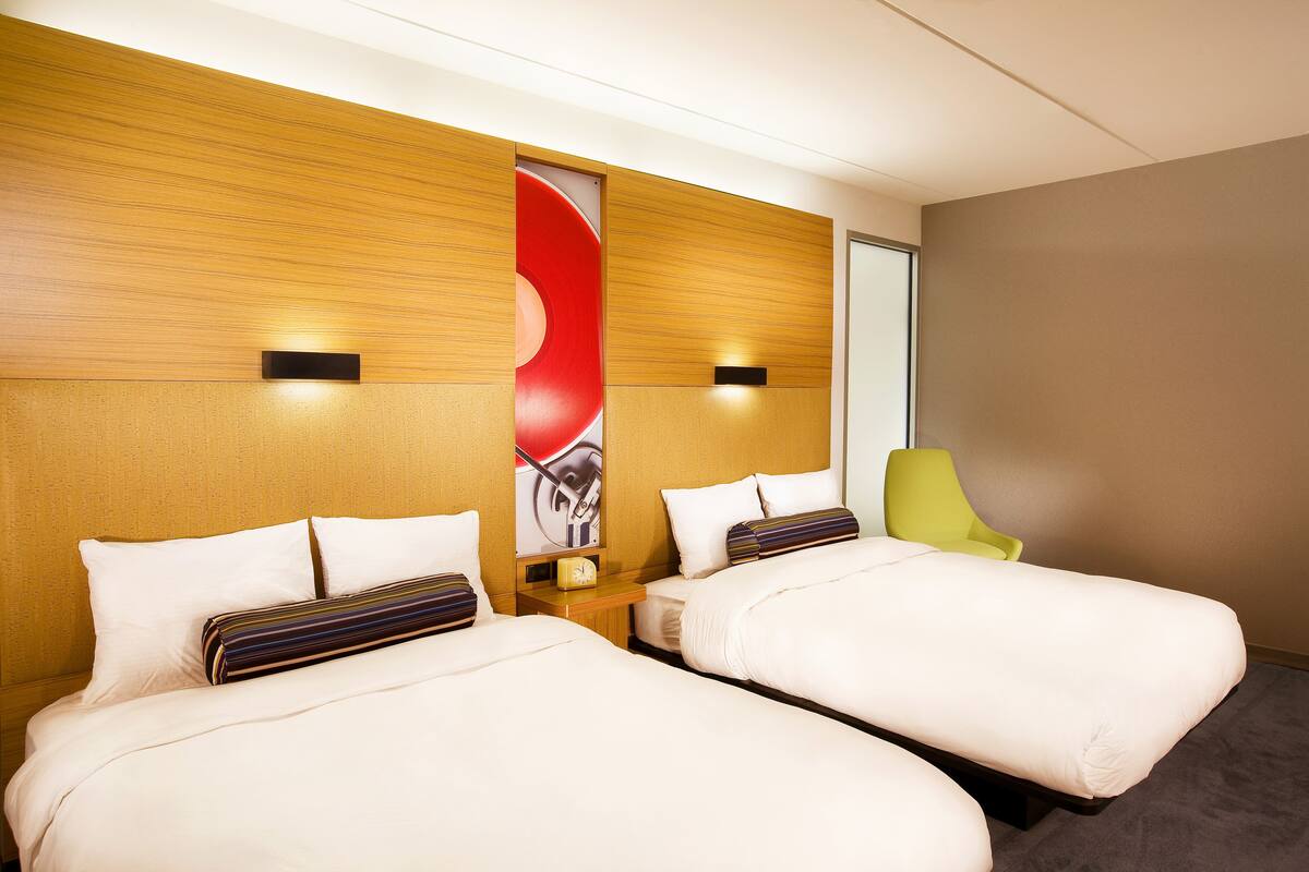 A hotel room with two beds, a wooden headboard, a red abstract artwork, a green chair, and a small table with a clock.