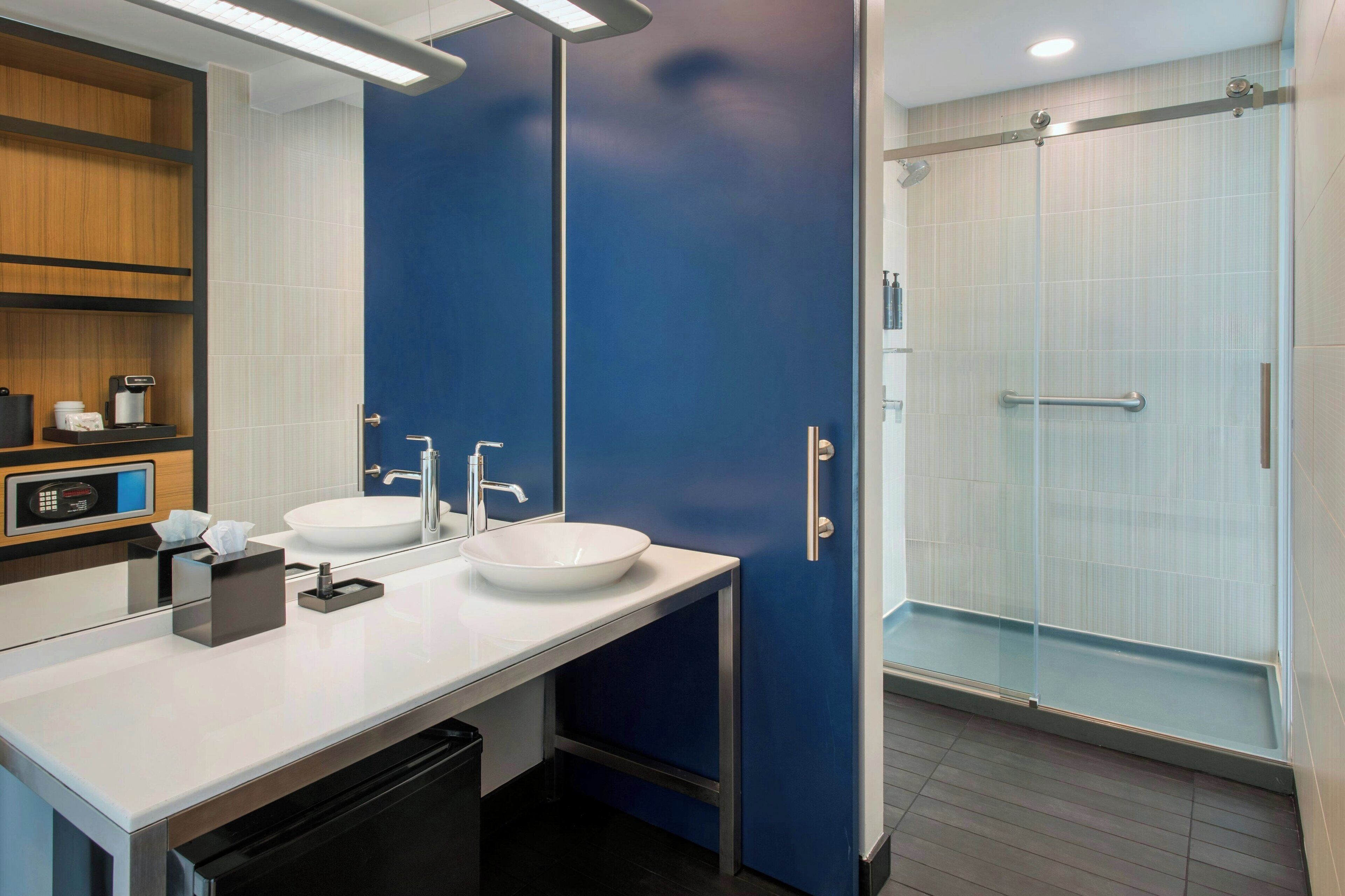 Un bany modern amb un lavabo blanc, un mirall, una dutxa amb porta de vidre i una prestatgeria de fusta amb productes de tocador.