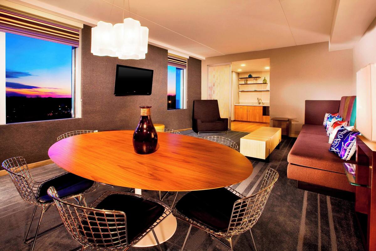 A modern hotel room with a round dining table, wireframe chairs, a sofa, a TV mounted on the wall, and a view of the city at sunset.