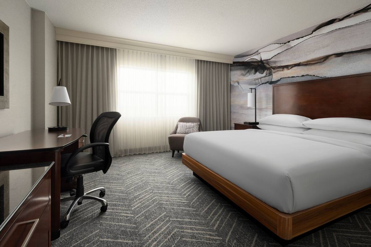 A hotel room with a bed, desk, chair, and a large window with curtains.