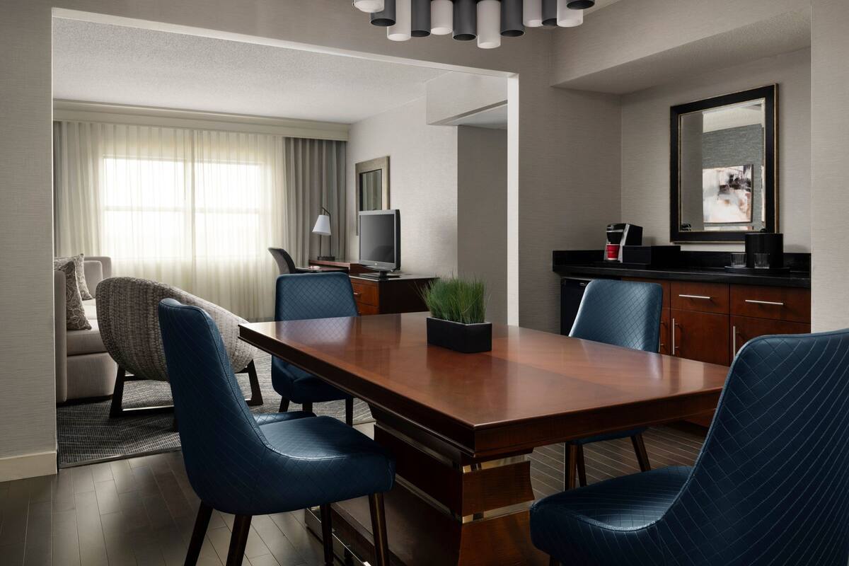 A modern hotel room with a wooden dining table, blue upholstered chairs, a television, a coffee machine, and a large mirror.