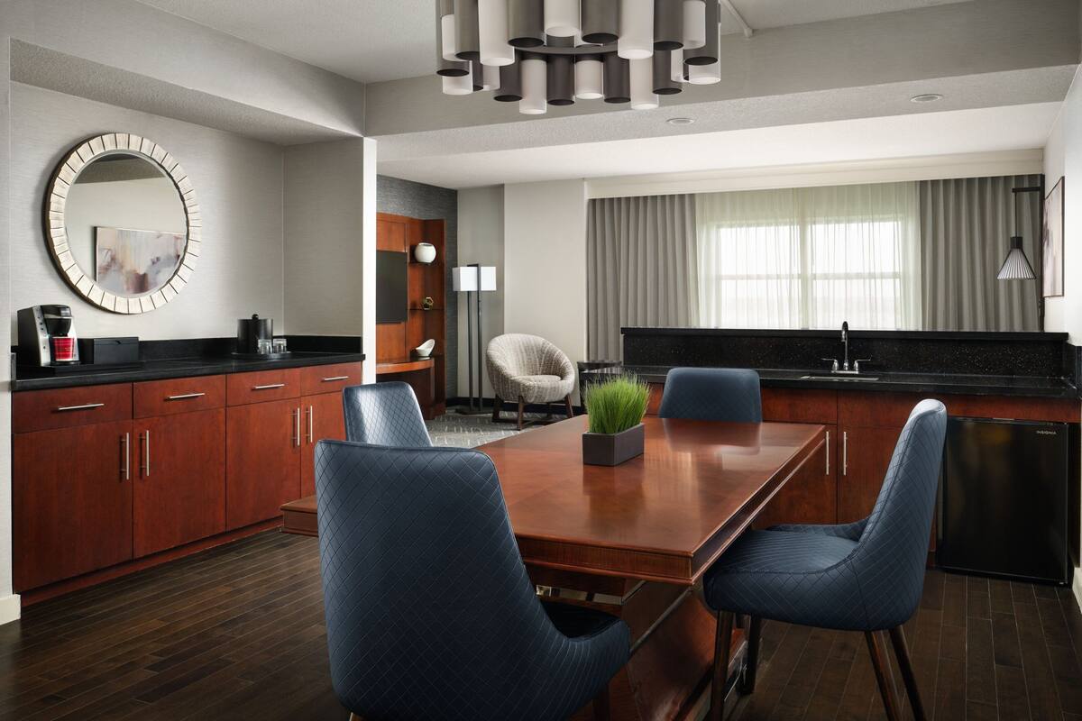 A modern kitchen with a dining area, featuring a wooden table, blue chairs, a coffee machine, and a refrigerator.
