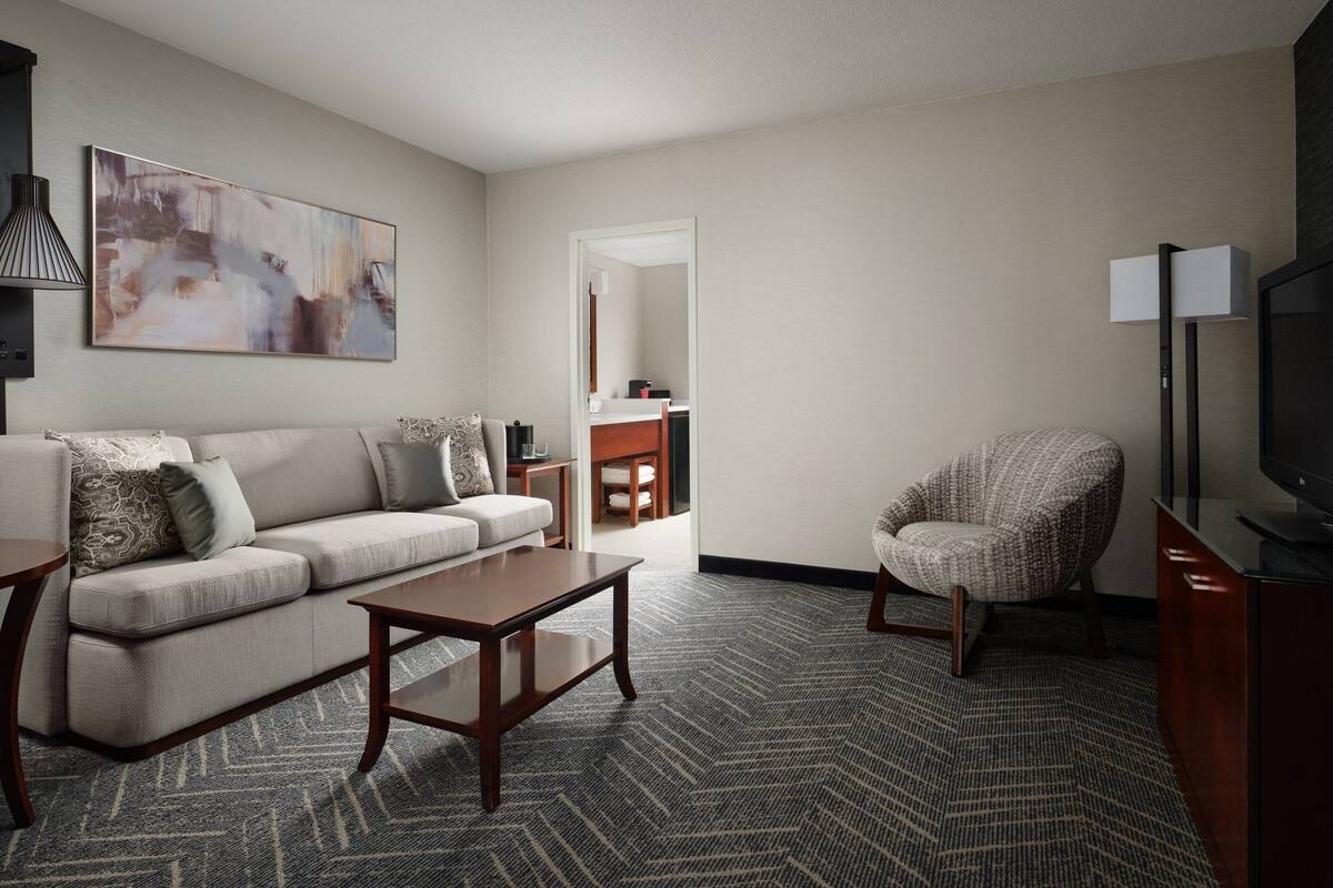 A modern hotel room with a sofa, armchair, coffee table, and a flat-screen TV.