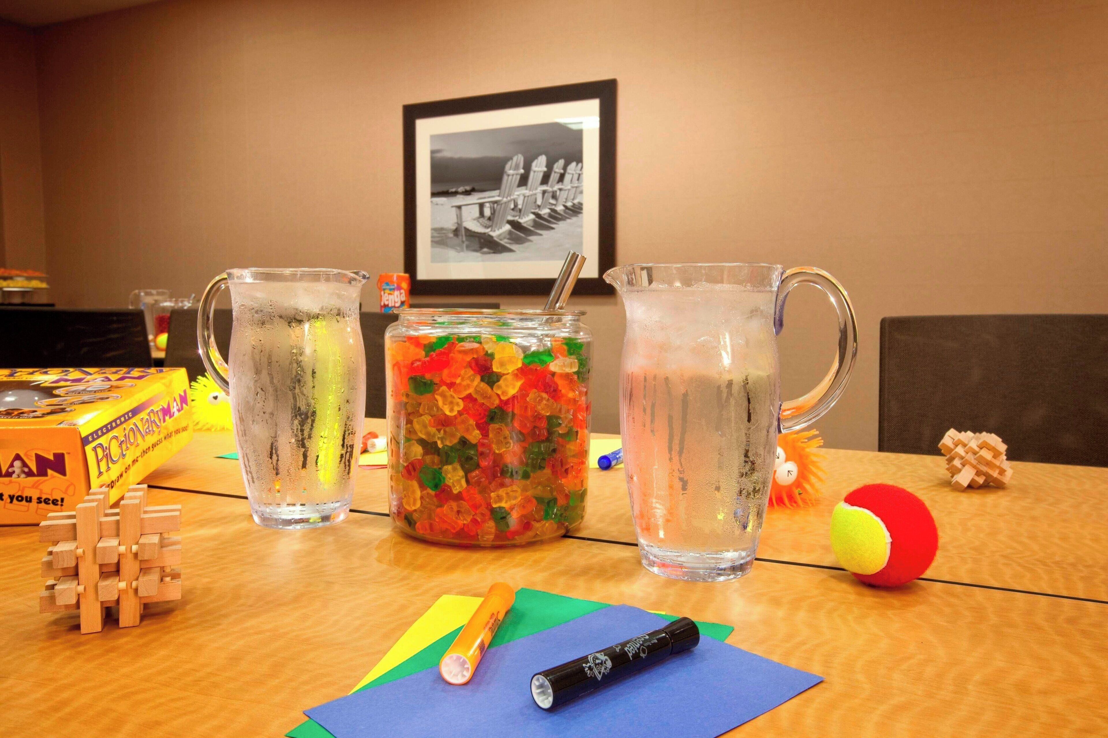 A table with two pitchers of water, a jar of gummy bears, a wooden puzzle, and a framed picture on the wall.