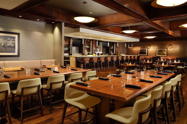 A restaurant with multiple long wooden tables and chairs, a bar area with a TV, and framed pictures on the walls.