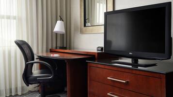 A hotel room with a wooden desk, a black office chair, a flat-screen TV, a lamp, and a mirror.