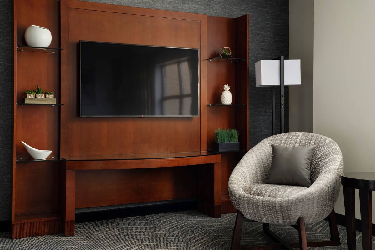 A modern living room with a large flat-screen TV mounted on a wooden cabinet, a comfortable armchair, and a floor lamp.