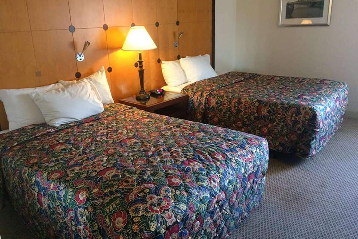 A hotel room with two beds, each with floral-patterned bedding, a wooden headboard, a nightstand with a lamp, and a framed picture on the wall.