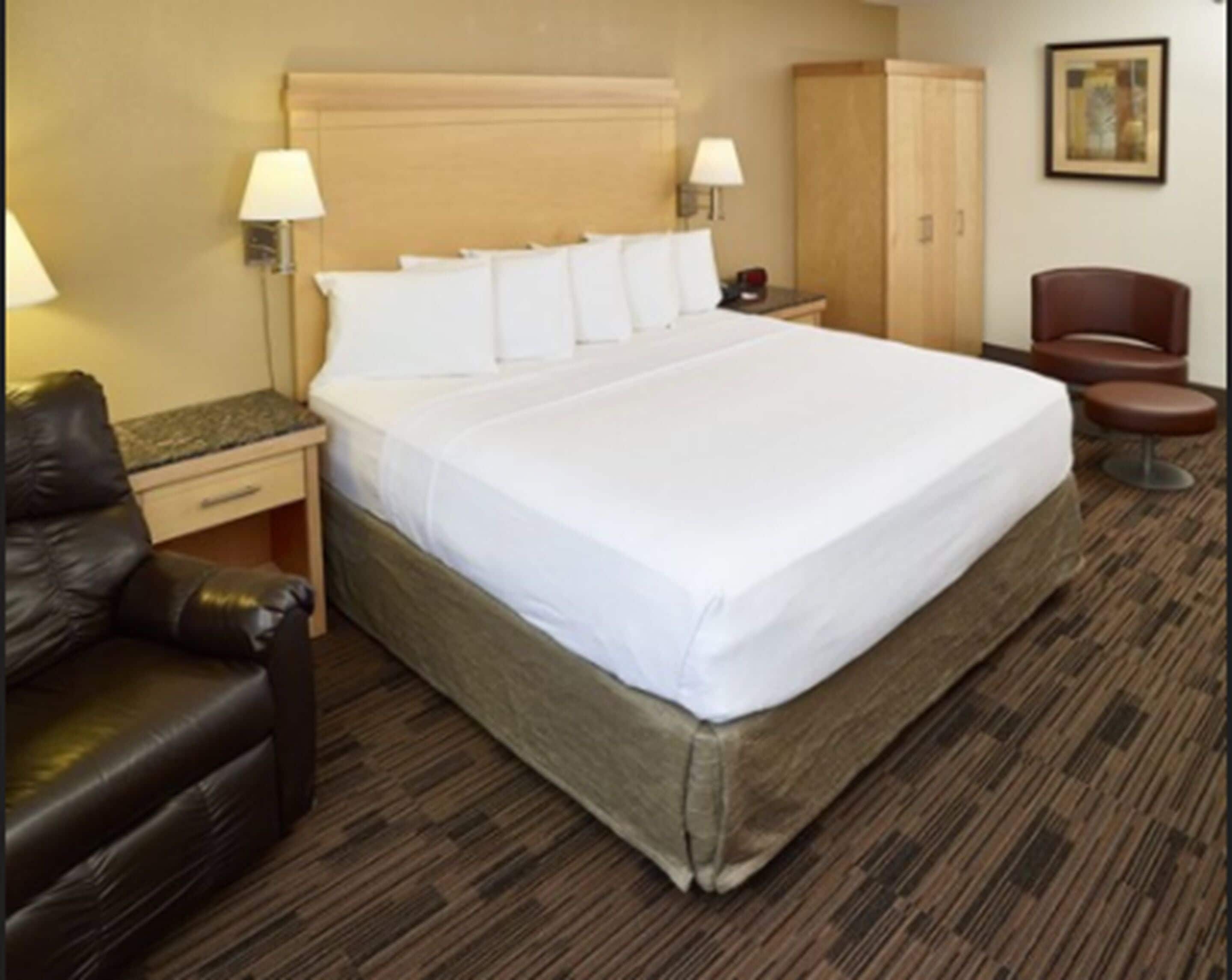 Una habitación de hotel con una cama grande, un cabecero de madera, dos lámparas de noche, un armario de madera, un sillón de cuero y una pequeña mesa redonda.