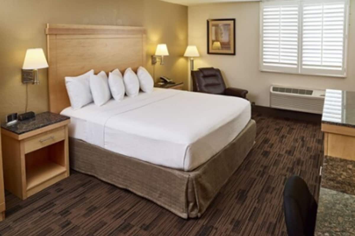 A hotel room with a large bed, two bedside tables with lamps, a chair, a desk, and a window with blinds.