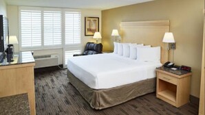 Habitación de hotel con una cama grande, dos mesitas de noche con lámparas, un escritorio con ordenador, una silla y un reposapiés marrón.