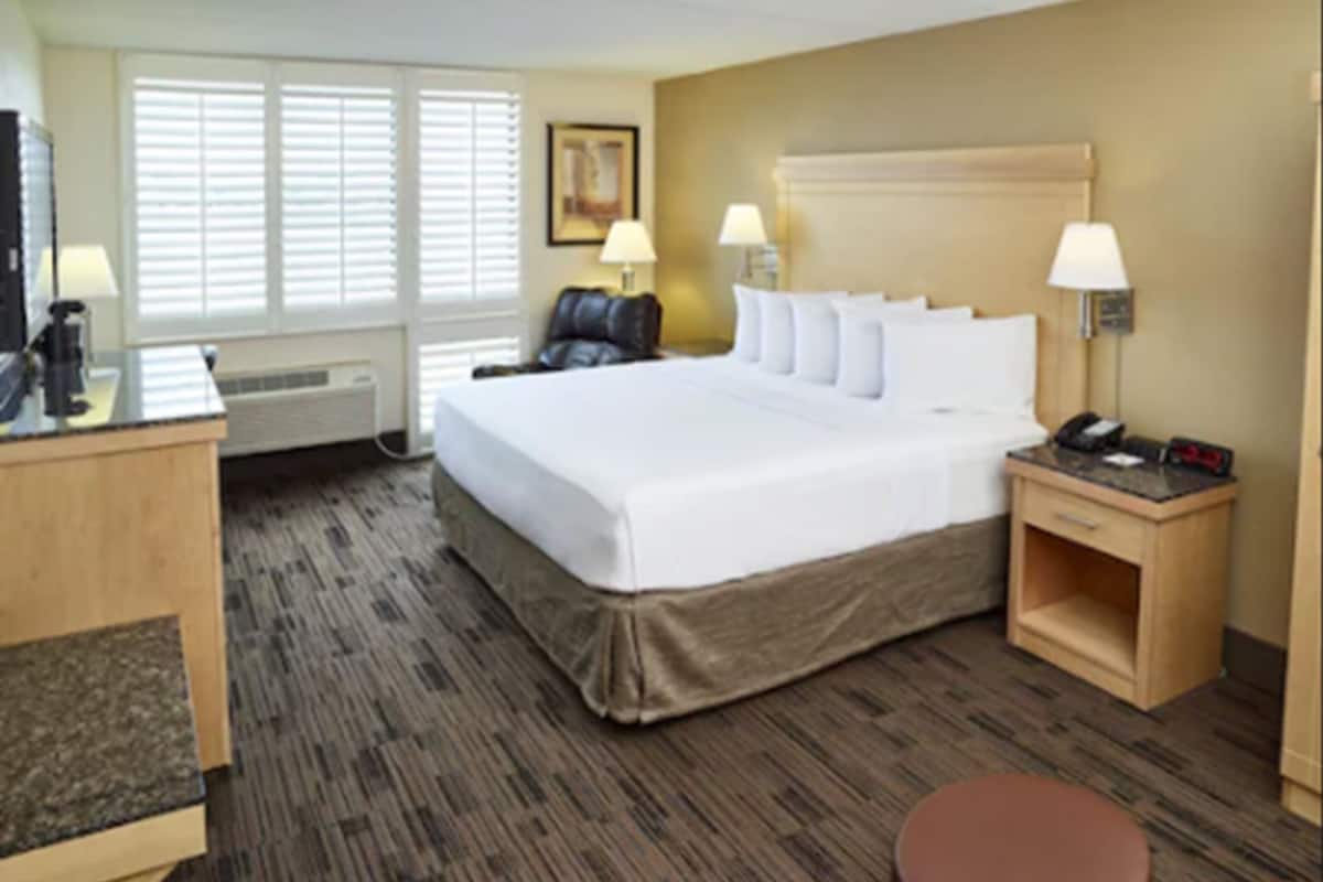A hotel room with a large bed, two bedside tables with lamps, a desk with a computer, a chair, and a brown ottoman.