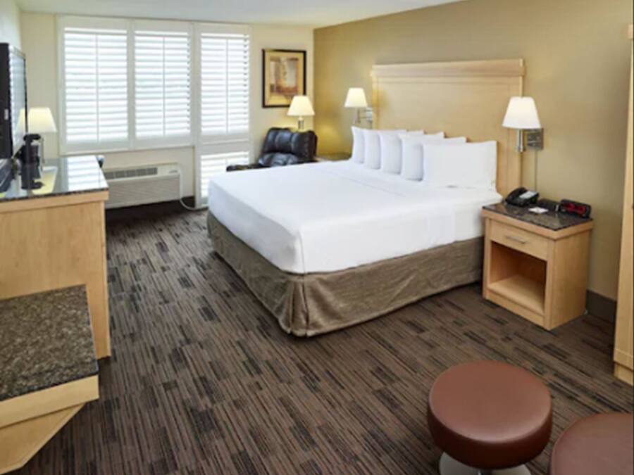 Une chambre d’hôtel avec un grand lit, deux tables de chevet avec des lampes, un bureau avec un ordinateur, une chaise et un pouf marron.