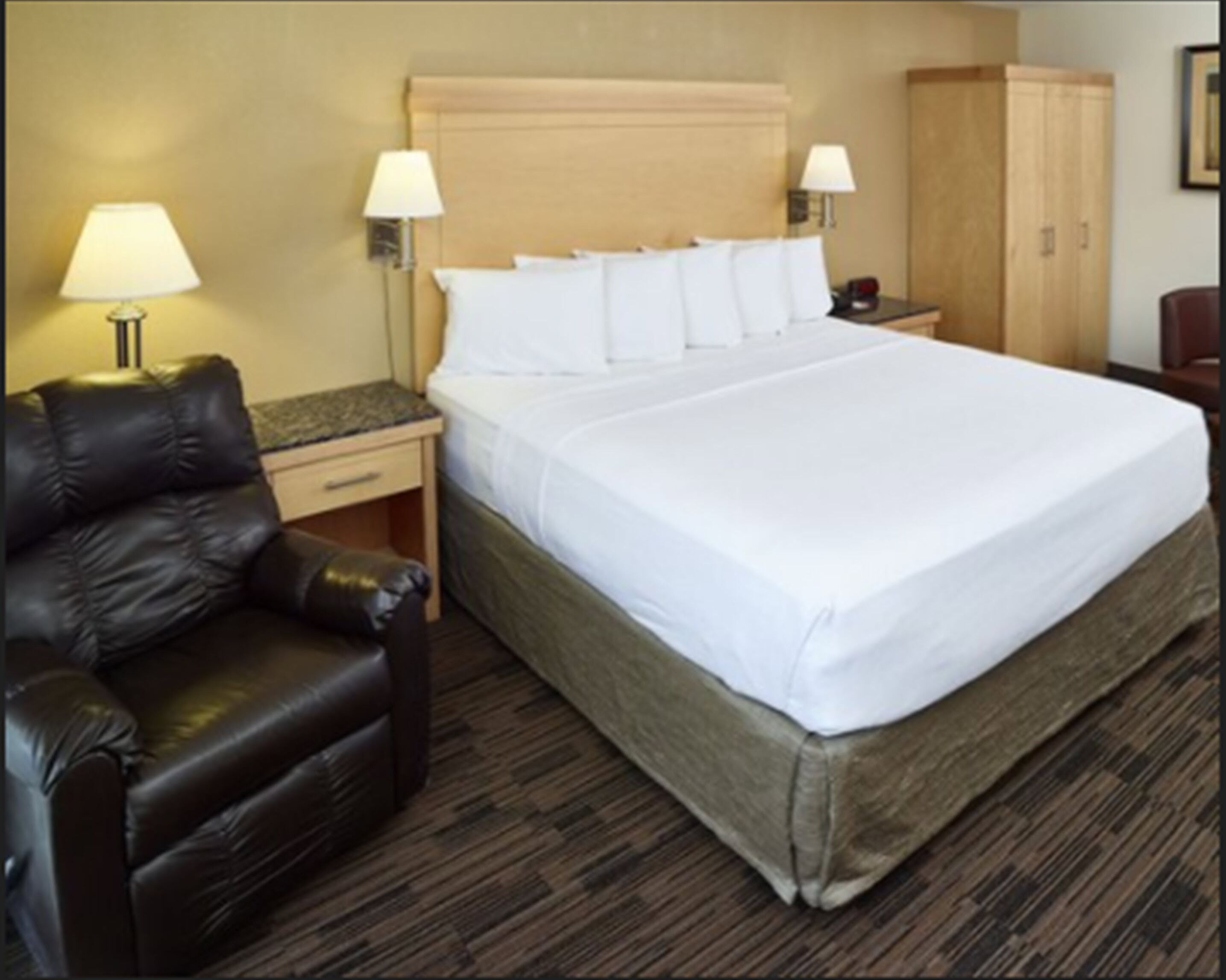 A hotel room with a large bed, two bedside tables, a nightstand, a leather armchair, and a wardrobe.