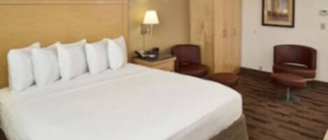 A hotel room with a large bed, a wooden headboard, a desk with a lamp, a chair, and a wardrobe.