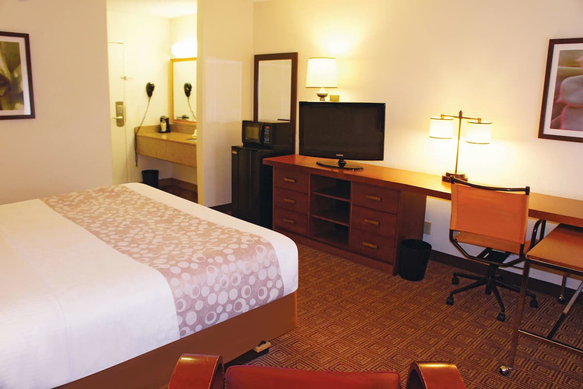 A hotel room with a bed, a desk with a television, a chair, a desk lamp, a mirror, and a small trash bin.