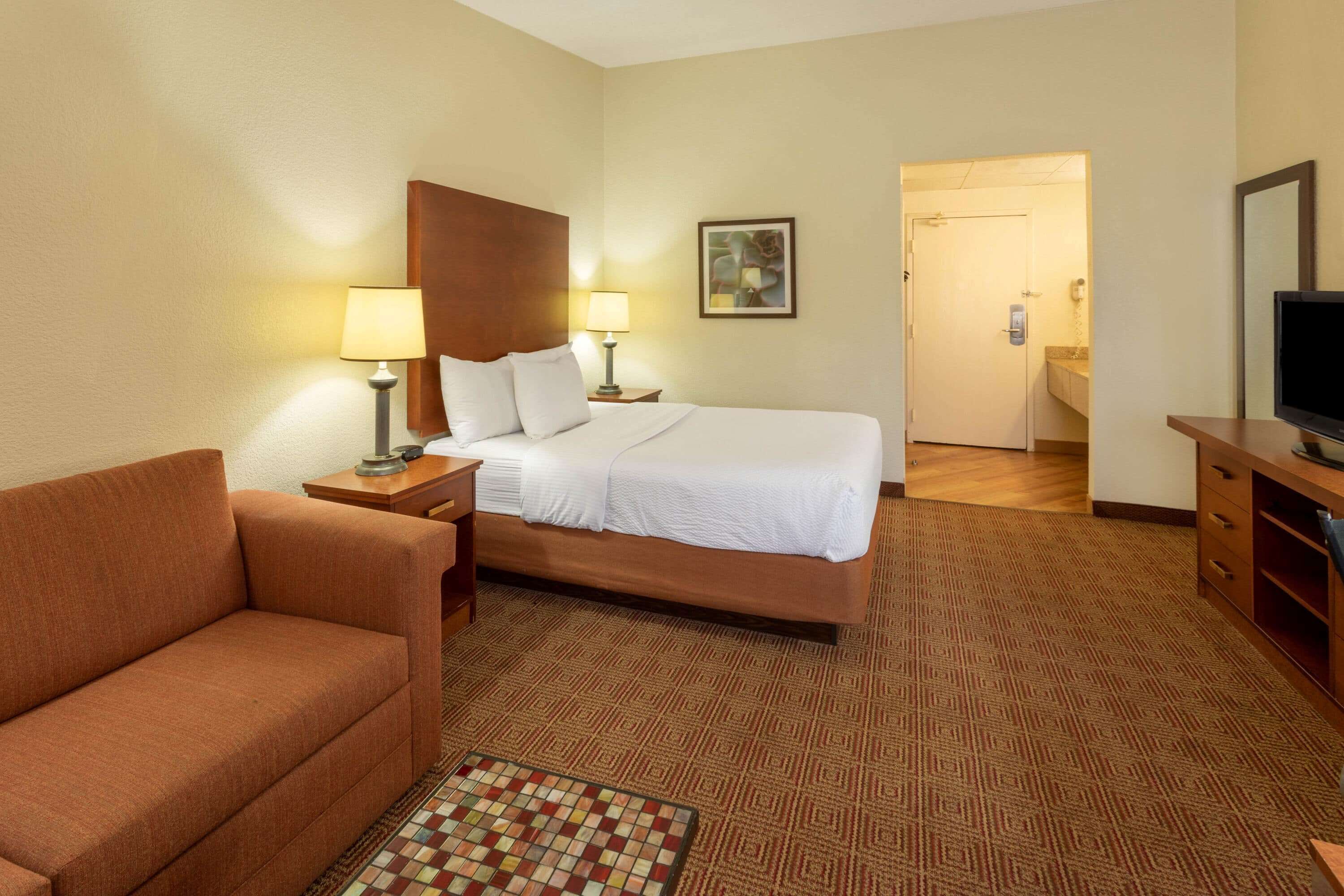 Habitación de hotel con cama, sofá, televisor y baño a la vista a través de una puerta abierta.