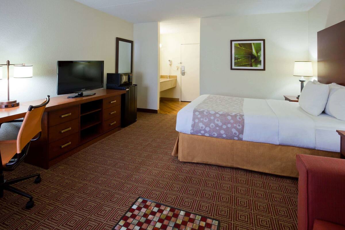 A hotel room with a bed, a desk with a television, a chair, a dresser, and a bathroom visible through an open door.