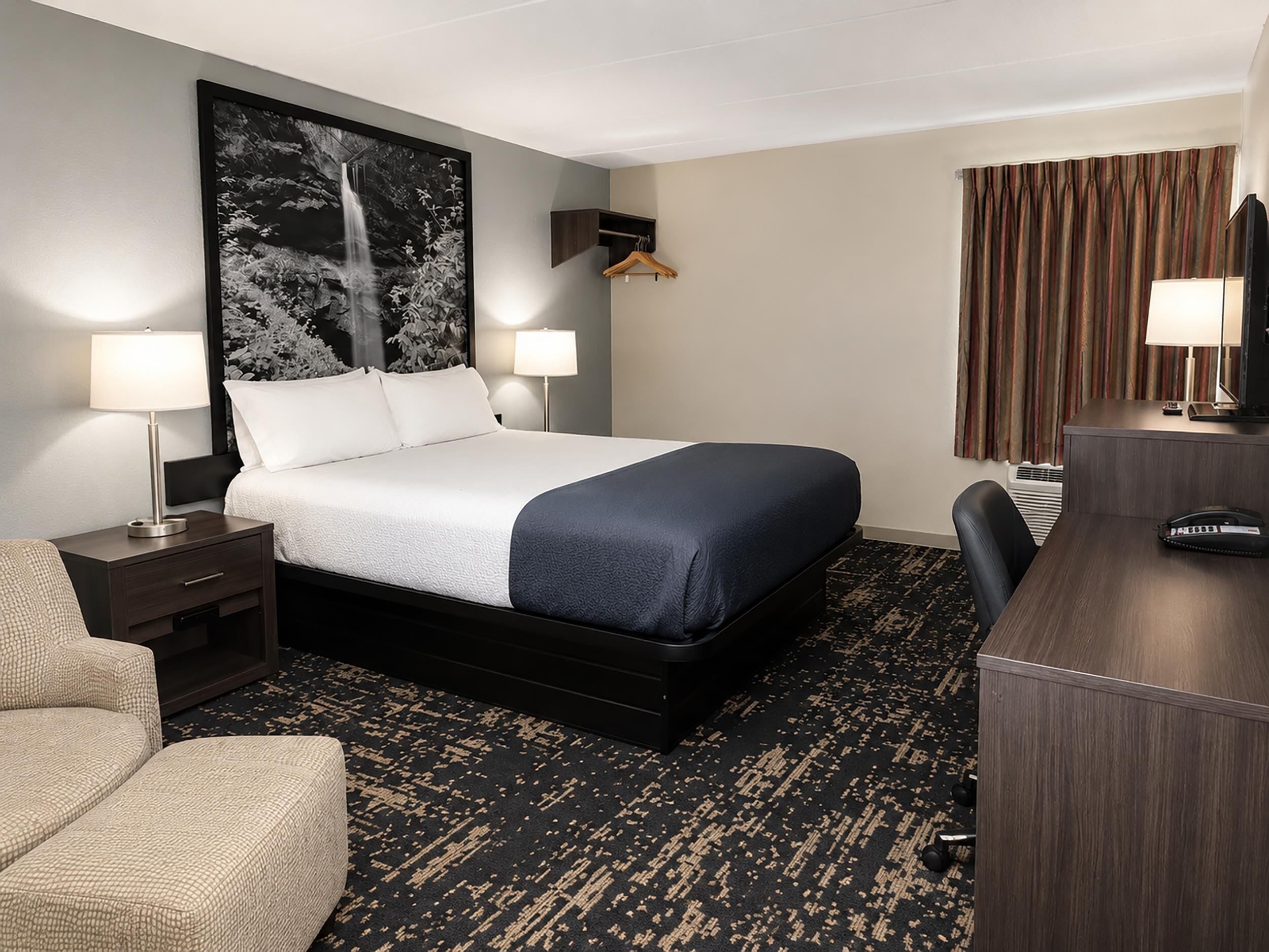 Habitación de hotel con una cama, dos lámparas de noche, un escritorio con teléfono, una silla y un cuadro abstracto grande en la pared.