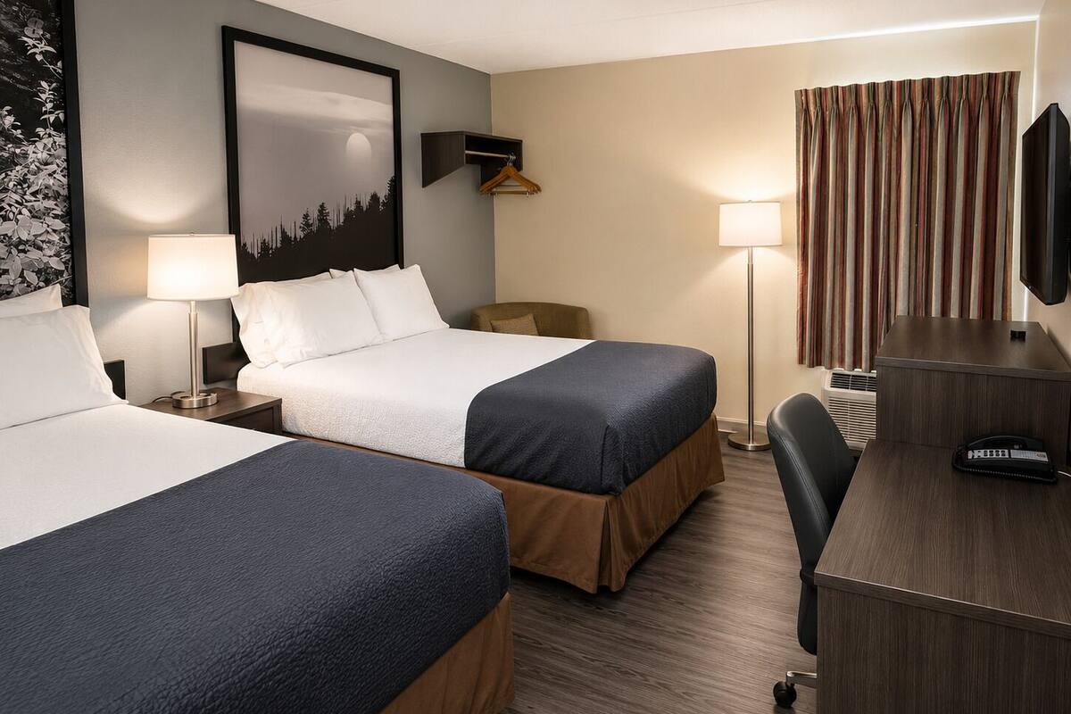 Ein Hotelzimmer mit zwei Betten, einem Schreibtisch, einem Sessel, einem Fernseher, einer Lampe und einem gerahmten Bild an der Wand.